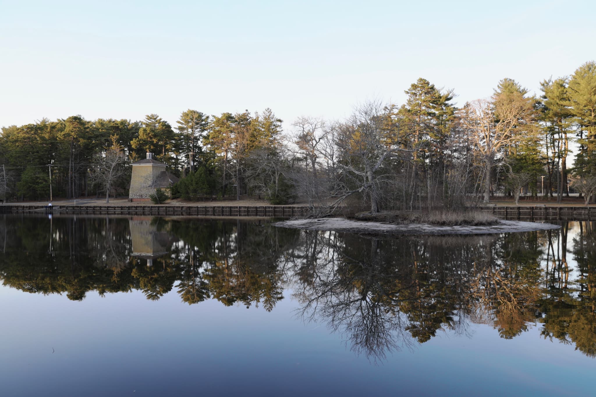 A calm lake reflecting a line of trees and a small structure, with a clear sky above